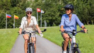 Zwei Frauen auf E-Bikes bei sommerlicher Tour mit Flaggen von Liechtenstein, Schweiz und Österreich im Hintergrund